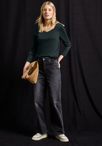 Tmavě zelený top s dlouhým rukávem, s výstřihem do V, spárovaný s černými džínami s vysokým pasem a světlými teniskami. Drží béžovou suédovou tašku. Jednoduchý design, ležérní vzhled.