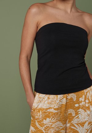 Vrouw met een zwarte top zonder bandjes en mosterdgele broek met witte bloemenpatronen, staand tegen een groene achtergrond.