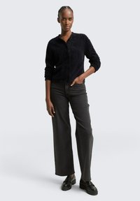 Schwarze, flauschige Strickjacke mit Rundhalsausschnitt und Knöpfen, kombiniert mit hochgeschnittenen, dunkelgrauen, weiten Jeans und schwarzen Plateau-Schuhen.