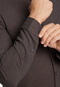 Camicia button-up marrone scuro con un tessuto testurizzato. Presenta un polsino con un singolo bottone e una vestibilità su misura. I bottoni sono dello stesso colore.