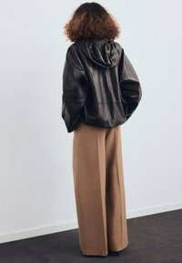 Giacca nera in pelle con cappuccio dal taglio rilassato, abbinata a pantaloni beige larghi, che mettono in risalto linee pulite e un design minimalista.