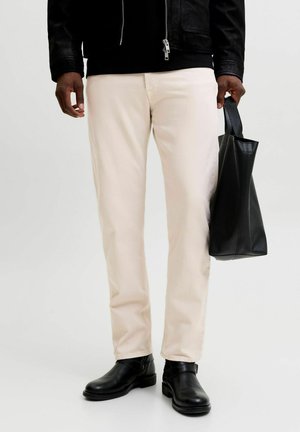 Homme portant un pantalon beige, des bottes noires et une veste noire tenant un sac fourre-tout en cuir noir devant un fond uni.