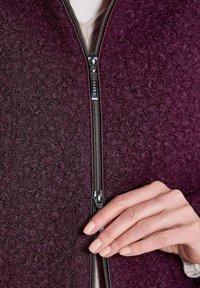 Strickjacke in Burgunderrot mit einer strukturierten Wolloberfläche und einem silbernen Reißverschluss, ergänzt durch eine Hand, die sanft den Reißverschlussgriff hält.