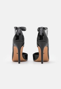 Svarta lackade högklackade skor med öppen baksida, spetsig tå och ankelremmar, med en dekorativ kedja som accent.