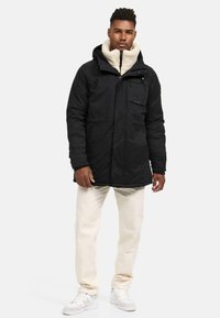 Parka negra con líneas, con capucha, que presenta un cierre de cremallera, cordón ajustable y un bolsillo en el pecho; llevada sobre un forro polar crema y pantalones beige.