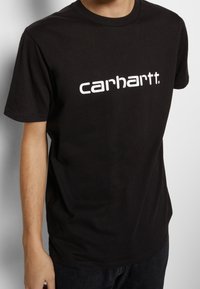 Schwarzes Baumwoll-T-Shirt mit kurzen Ärmeln, das ein auffälliges weißes "Carhartt."-Logo auf der Brust und einen runden Halsausschnitt aufweist.
