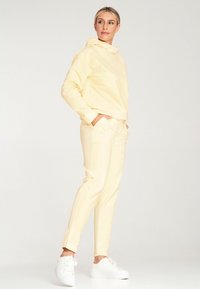 Żółta chusta z wysoką szyją i luźnym krojem, połączona z dopasowanymi żółtymi spodniami. Model nosi białe trampki. Gładka faktura.