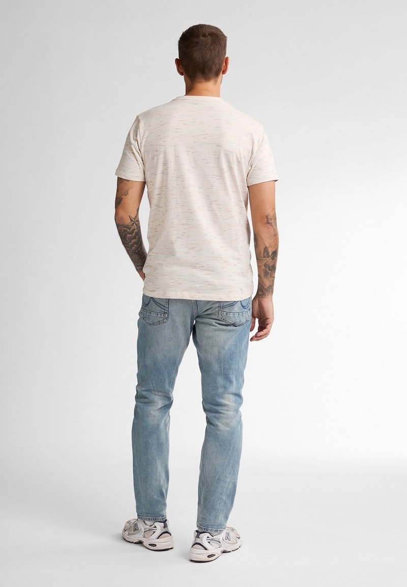 Mann mit Tätowierungen auf beiden Armen, der ein hell gesprenkeltes T-Shirt, blaue Jeans und weiße Sneakers trägt und vor einem weißen Hintergrund steht.