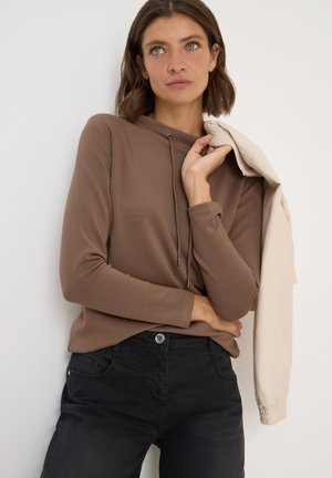 Bruine lange mouwen top met een capuchon en veters, gecombineerd met zwarte jeans. Beige jas hangt over één schouder.