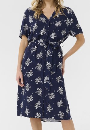 Kobieta w granatowej sukience na guziki z białym kwiatowym wzorem i paskiem związanym w talii, stojąca z jedną ręką na biodrze.