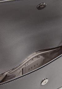 Šedý syntetický vnitřek kabelky s hladkou texturou, vybavený stříbrným cvokem a štítkem označeným "DKNY NEW YORK."