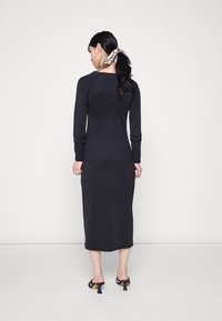 Vestido preto de mangas longas, com formato ajustado, textura canelada e comprimento até a metade da panturrilha. Apresenta um decote em U discreto e punhos dobrados.