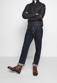 Giacca nera imbottita con colletto alto, con una cerniera arancione, abbinata a jeans in denim scuro e stivali marroni con lacci, arrotolati sui polsi.