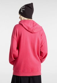 Roze hoodie met een relaxte pasvorm, ribbelboorden en -zoom. Draagt een zwarte beanie met witte grafische details.