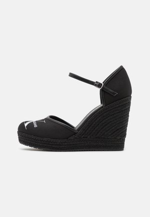 Espadrille de cuña negra con correa al tobillo y hebilla, con punta cerrada que presenta un logo impreso en blanco en la parte superior de tela.