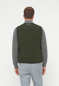 Roheline tikandiga vest kõrge kraega, millel on kaeluse juures logo, kantud halli pikkade varrukatega särk üle. Sile kangast tekstuur.