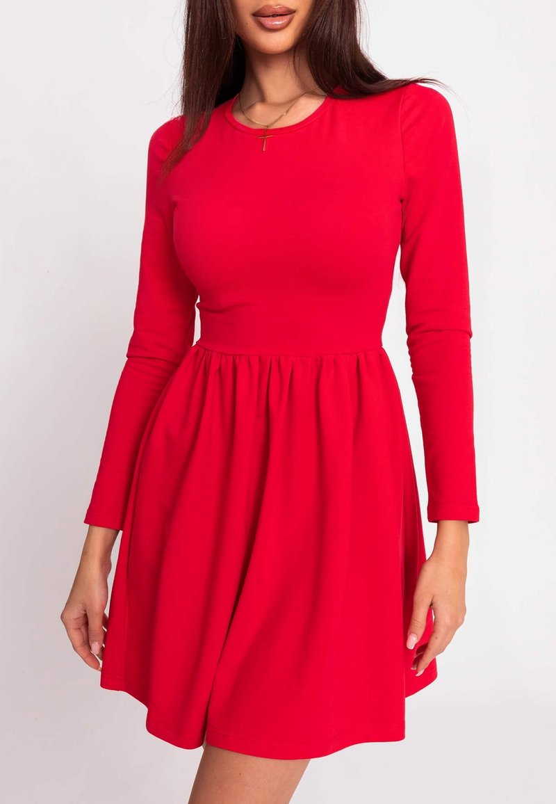 Femme portant une robe rouge cintrée à manches longues avec une taille marquée et un collier pendentif avec une croix.