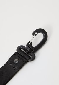Cinghia in nylon nera con una robusta clip in plastica nera, dotata di un gancio metallico e una texture liscia, cuciture dettagliate e un bottone decorativo.