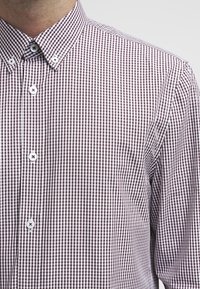 Mann trägt ein langärmliges, knöpfbares Hemd mit einem braun-weißen, klein karierten Muster und weißen Knöpfen.