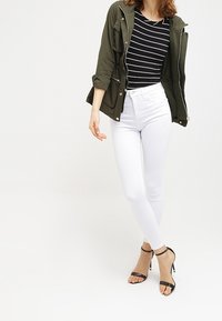 Kurtka oliwkowa, czarno-biała bluzka w paski, białe obcisłe dżinsy i czarne sandały na wysokim obcasie z paskami. Kurtka posiada zamek błyskawiczny i zatrzaski.