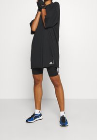 Camiseta deportiva negra de gran tamaño con mangas cortas, que presenta un logo en el dobladillo, combinada con pantalones cortos negros y zapatillas de running azules.