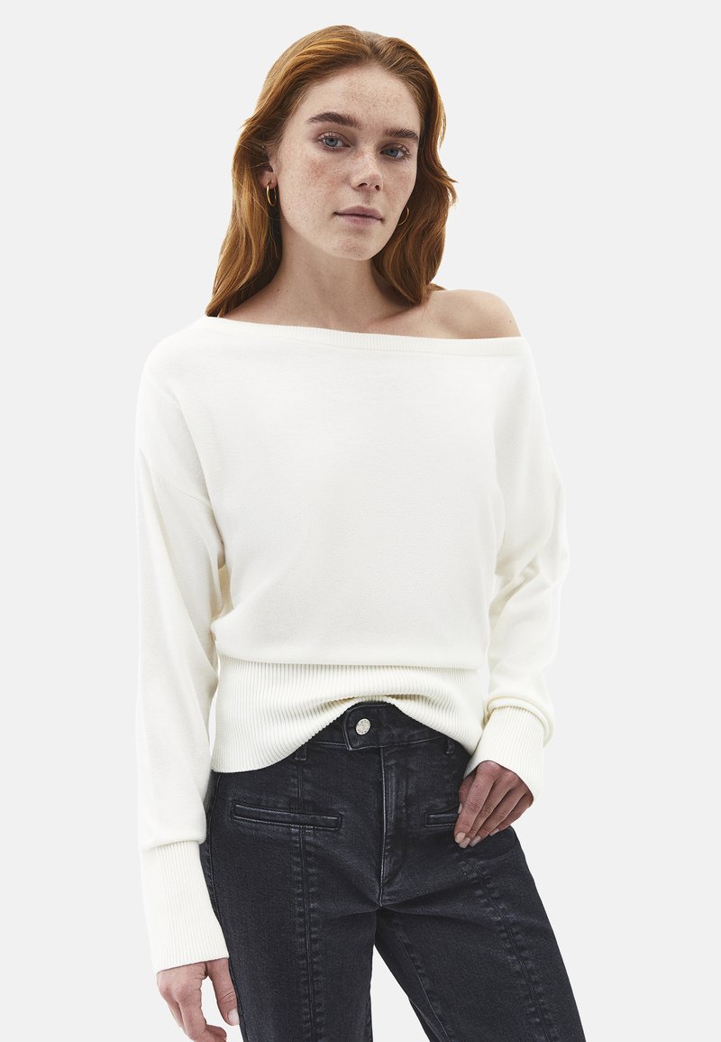 Valkoinen olkapäät paljastava neule, jossa pitkät hihat, ribbireunukset ja pehmeä materiaali, yhdistettynä mustiin farkkuihin, joissa on etutaskut.