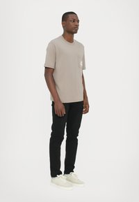 T-shirt beige a maniche corte e collo rotondo con tasca, abbinato a jeans neri e sneaker chiare. Tessuto morbido, vestibilità casual.