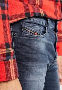 Mörkblå denimjeans med en smickrande passform, prydda med en liten röd och svart Diesel Industries-tag på framsidan av fickan, kombinerade med en röd rutig flanellskjorta.