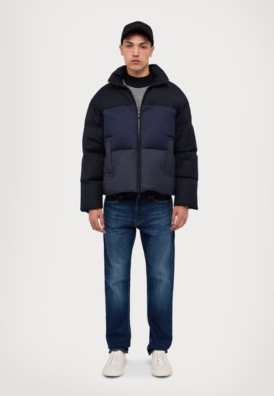 Puffer jacket in navy and dark blue, zippered front, elastic cuffs, worn over a striped sweater, paired with blue jeans and white sneakers.