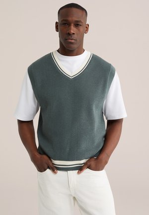 Mees kannab rohelist kootud varrukateta kampsunisalli, millel on valge V-kaelusega ääris, valge lühikeste varrukatega särgi kohal, käed taskutes, neutraalne taust.