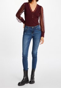 Bordeauxfarbenes Body mit V-Ausschnitt, Spitzenbesatz und durchsichtigen gepunkteten Puffärmeln, kombiniert mit blauen Skinny Jeans und schwarzen Knöchelschuhen.