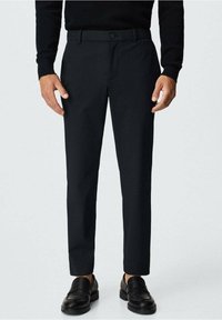 Černé kalhoty na míru, ve slim střihu, vyrobené z hladké látky. Mají zapínání na knoflík a boční kapsy, doplněné černými mokasíny.