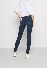 Jeans ajustados de mezclilla azul oscuro, con cintura alta, bolsillos traseros y una textura suave, combinados con una camiseta blanca holgada.