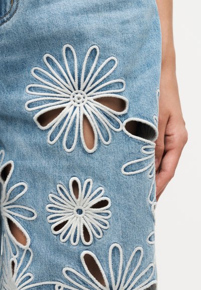 Ανοιχτό μπλε τζιν ύφασμα με μεγάλα κεντημένα λευκά μοτίβα μαργαρίτας που διαθέτουν κομμένα πέταλα, που δείχνουν το δέρμα από κάτω και ένα χέρι που ακουμπά κοντά.