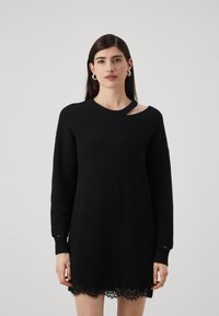Abito nero in maglia con un tessuto testurizzato, scollo a barchetta con dettaglio a taglio e orlo in pizzo. Maniche lunghe con polsini a costine.