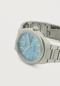 Timex kvarcinis laikrodis su šviesiai mėlynu ciferblatu, sidabriniais valandų žymekliais, nerūdijančio plieno korpusu ir apyranke, bei datos rodymu prie 3 valandos.