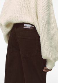 Maglione lavorato a maglia color crema con una vestibilità rilassata, indossato sopra pantaloni marrone scuro con una toppa in pelle e due tasche posteriori.