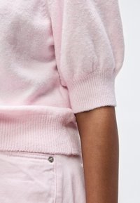 Krupni plan osobe koja nosi svijetloružičasti mekani džemper s napuhanih rukava i svijetloružičaste hlače s vidljivim metalnim gumbom.