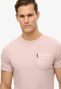 Camiseta de manga corta rosa claro, hecha de una tela suave, con un bolsillo en el pecho que presenta una pequeña etiqueta de la marca. Cuello redondo y corte relajado.