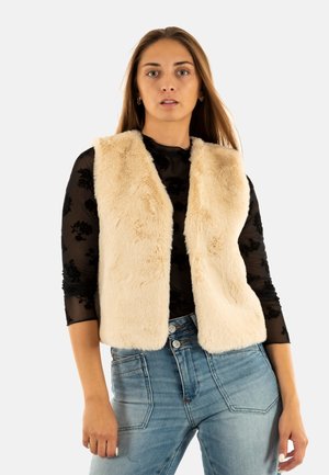 Chaleco de piel sintética en crema, con una textura suave, frente abierto y sin cierres, llevado sobre una blusa de malla negra con estampado floral y jeans de talle alto azules.