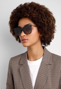 Black square sunglasses with thick frames, featuring a subtle logo detail. The model wears a checked blazer over a white shirt.