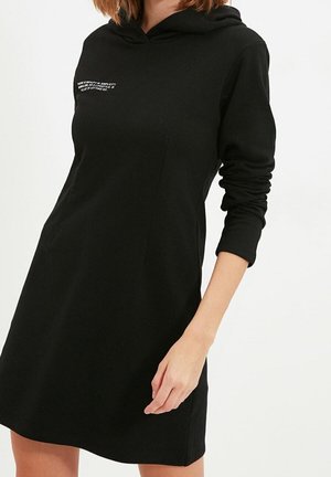 Zwarte hoodie jurk met lange mouwen, voorzien van een zak aan de voorkant en geborduurde tekst boven de linkerborst. Gemaakt van een zachte, rekbare stof.