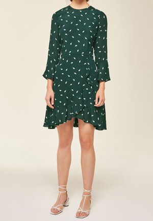 Robe verte avec des motifs de feuilles blanches, manches longues avec des poignets évasés,Ourlet en volants, fabriquée dans un tissu léger. Portée avec des sandales blanches à brides.