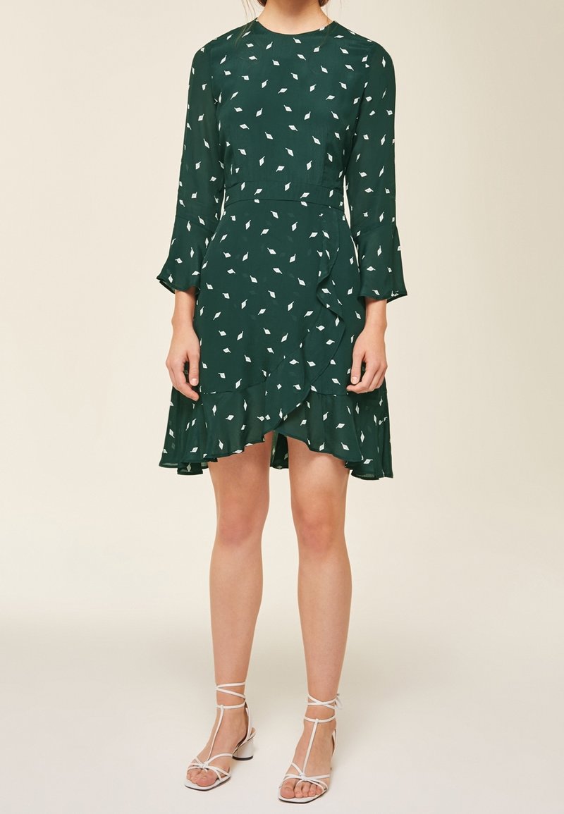 Vestido verde con patrones de hojas blancas, mangas largas con puños acampanados, dobladillo con volantes, hecho de una tela ligera. Llevado con sandalias blancas de tiras.