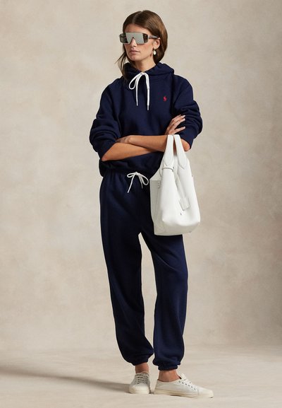 Γυναίκα με μπλε σκούρο φούτερ με κουκούλα και φόρμες, φοράει μεγάλα γυαλιά ηλίου, λευκά αθλητικά παπούτσια, κρατώντας μια λευκή δερμάτινη τσάντα ώμου, στέκεται με σταυρωμένα τα χέρια.