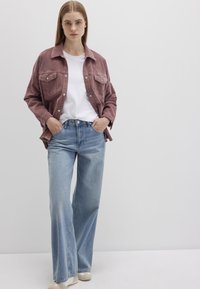 Rosa Cordjacke mit doppelten Brusttaschen über einem weißen T-Shirt, kombiniert mit hellblauen weiten Jeans und beigen Schuhen.