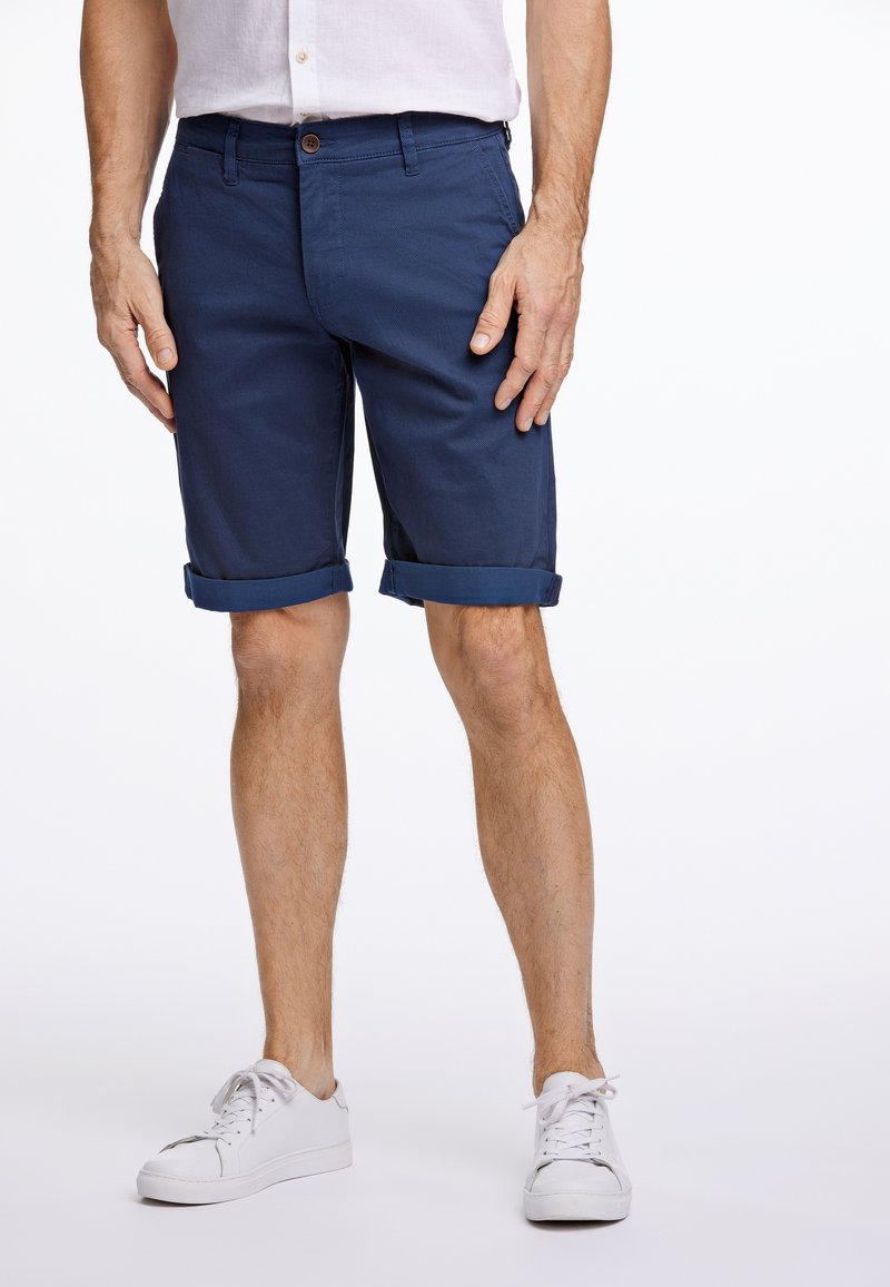 Mann trägt marineblaue, hochgekrempelte Shorts, weiße Sneakers und ein helles Kurzarmhemd und steht vor einem weißen Hintergrund.
