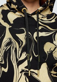Sudadera negra con un patrón abstracto dorado, con cordones negros y detalles de metal dorado. La tela tiene una textura suave.