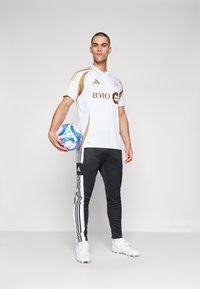 Camiseta de fútbol blanca con acentos dorados, pantalones atléticos negros con rayas blancas, sosteniendo un balón de fútbol multicolor, de pie frente a un fondo gris.