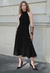 Crna halter haljina bez rukava s lepršavom suknjom, napravljena od glatke tkanine; uparena s ljubičastim štiklama i prozirnim smešnim akcentima na rukama.